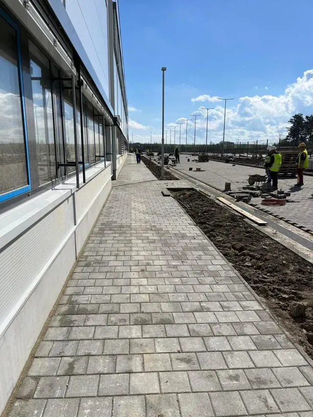 Téglakő járda épülő irodaház homlokzata mentén, háttérben parkolóépítéssel